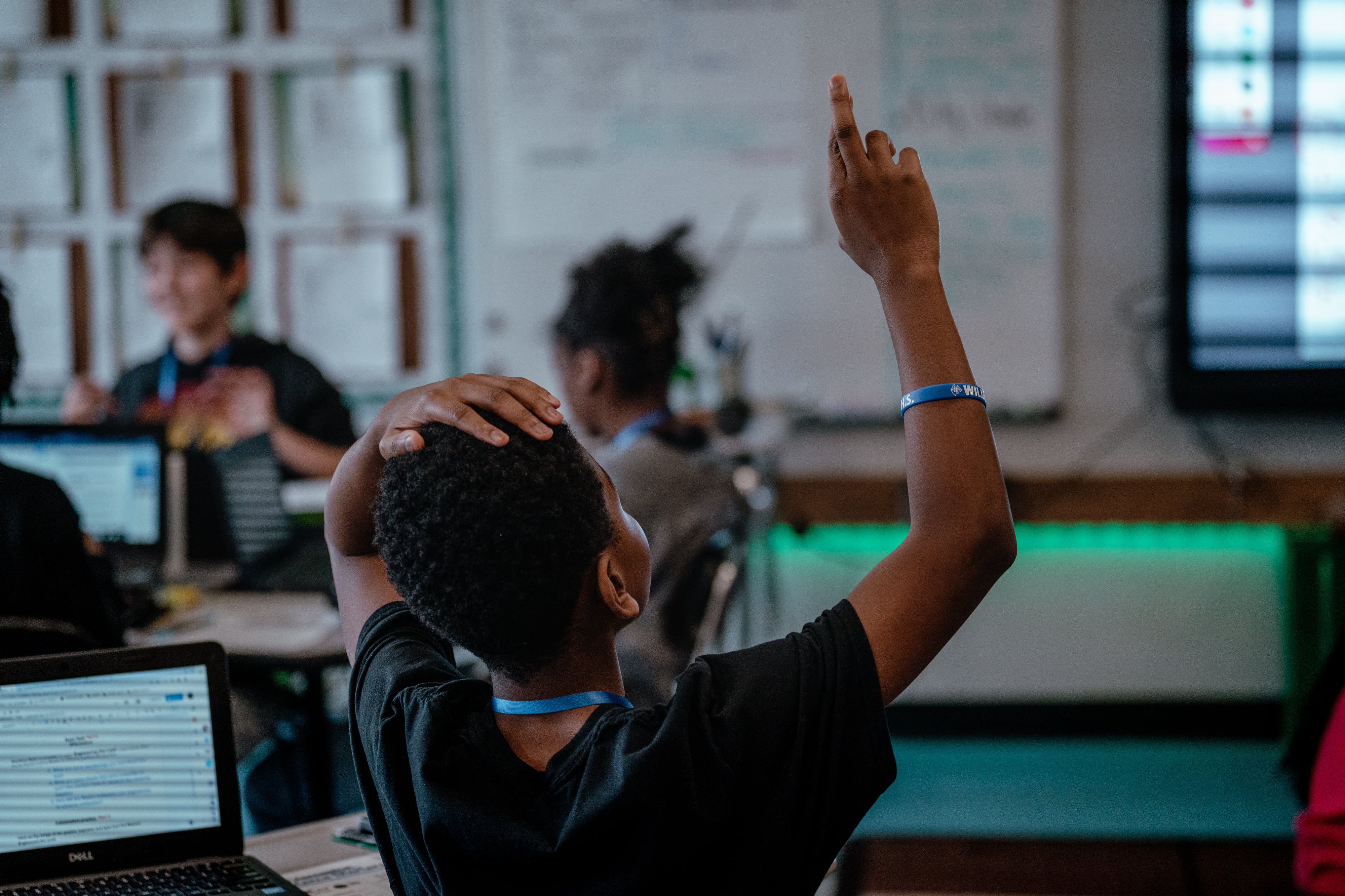 A back view of a student raising their hand in a classroom with other students in the background.