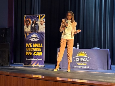 A Black woman in business clothes stands and speaks on stage with two banners in the background.