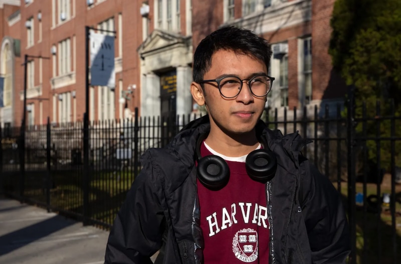 A boy with glasses wearing a black jacket over a Harvard sweatshirt and headphones around his neck stands in front of a school.