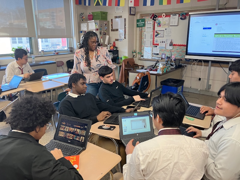 A Black teacher with long dark hair stands next to a group of high school students sitting at school desks working on their laptops in a classroom.