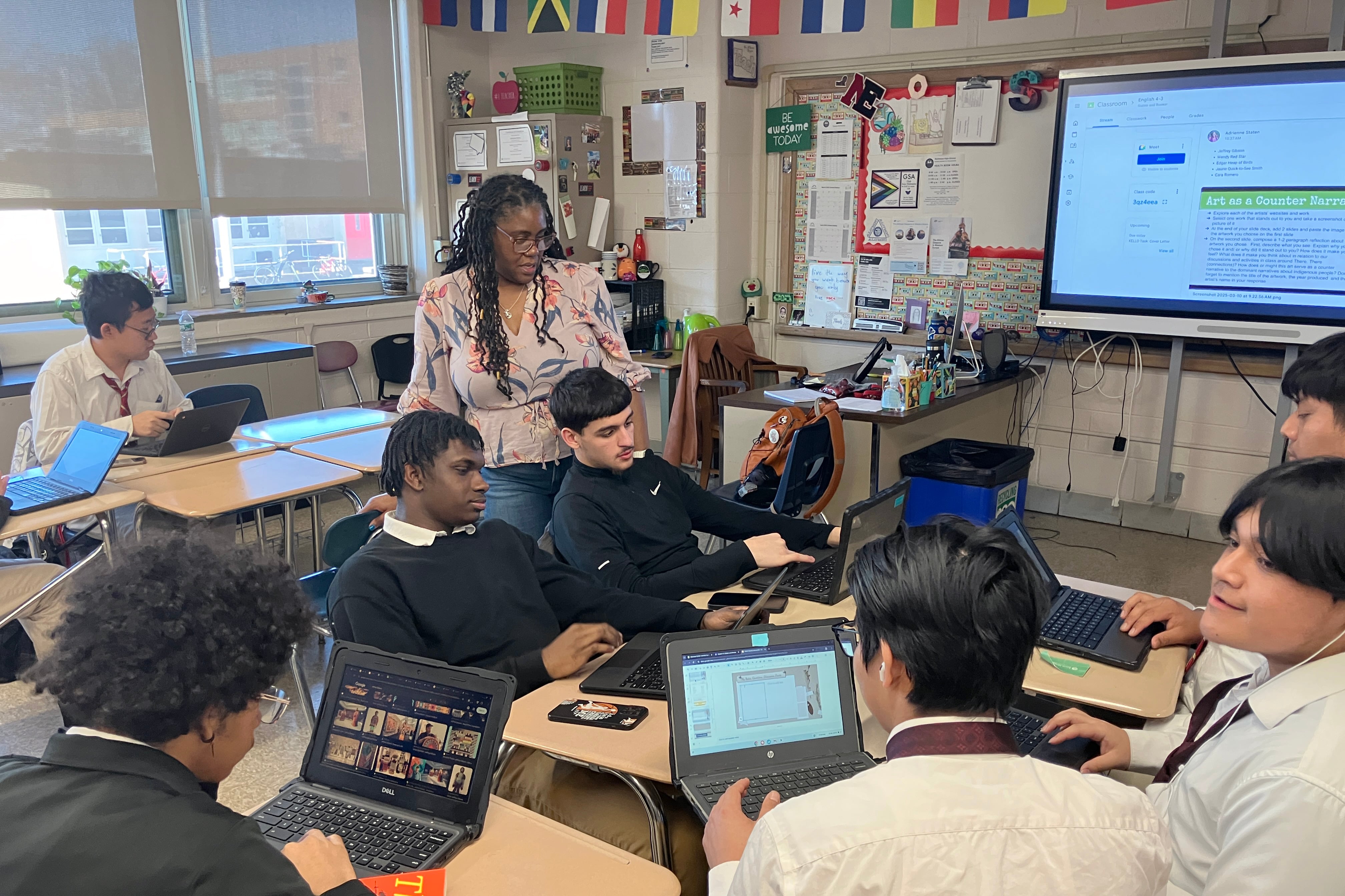 A Black teacher with long dark hair stands next to a group of high school students sitting at school desks working on their laptops in a classroom.