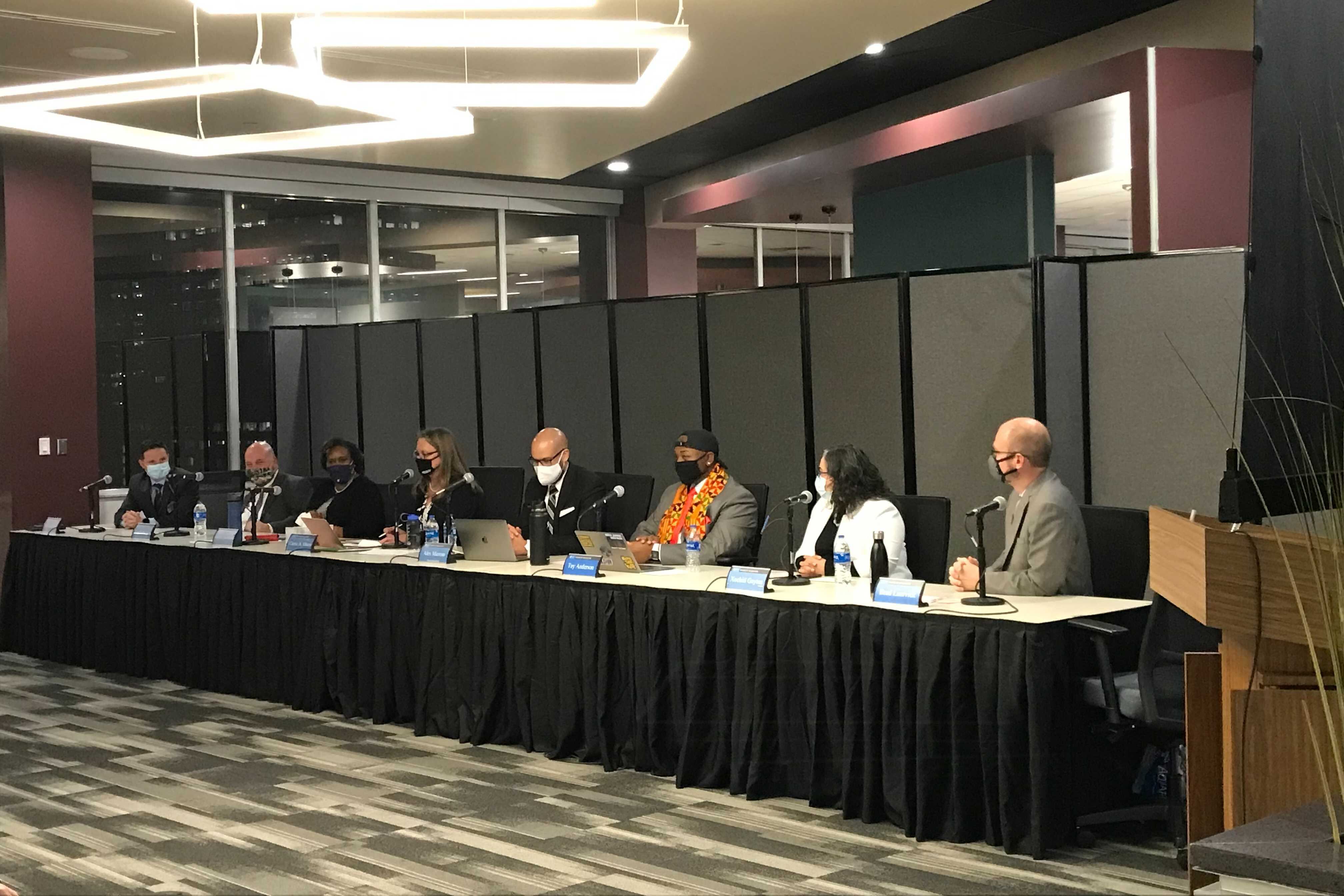 Men and women in business attire and masks sit at a long table in a conference room. They have microphones and name plates in front of them.