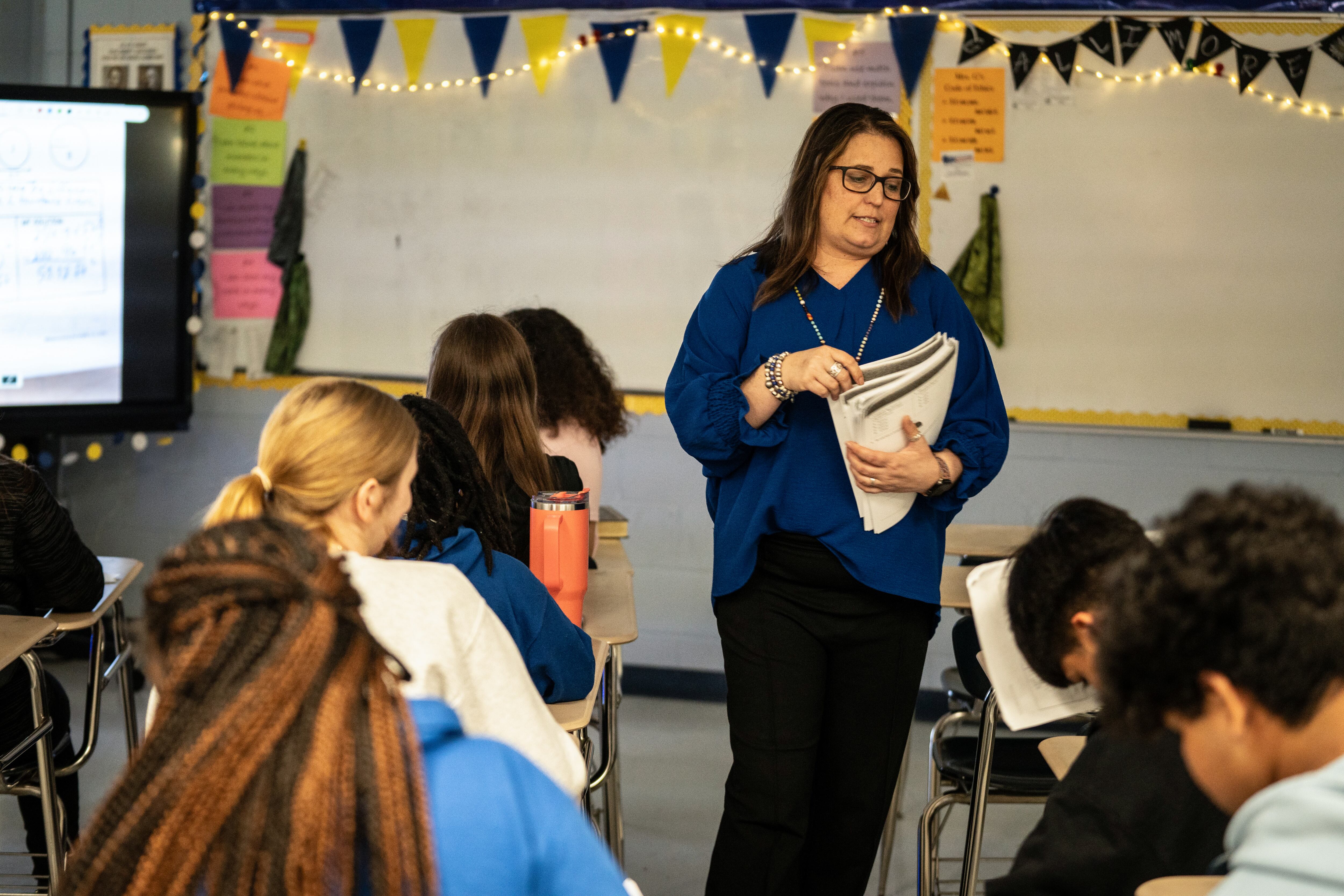 A woman with a blue top and glasses holds a stack of papers standing among students at desks.