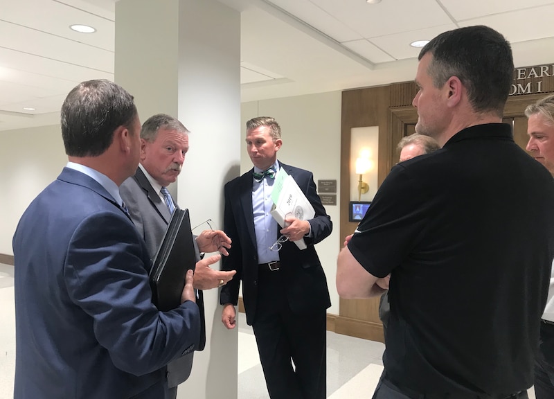 A group of five or more men in suits stand in a circle talking with each other in a hallway.