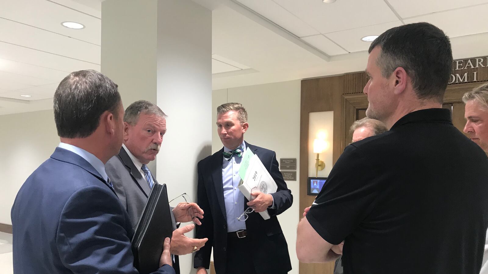 A group of five or more men in suits stand in a circle talking with each other in a hallway.