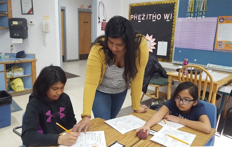 A photograph of a woman standing between two young students sitting at a table in a a classroom.