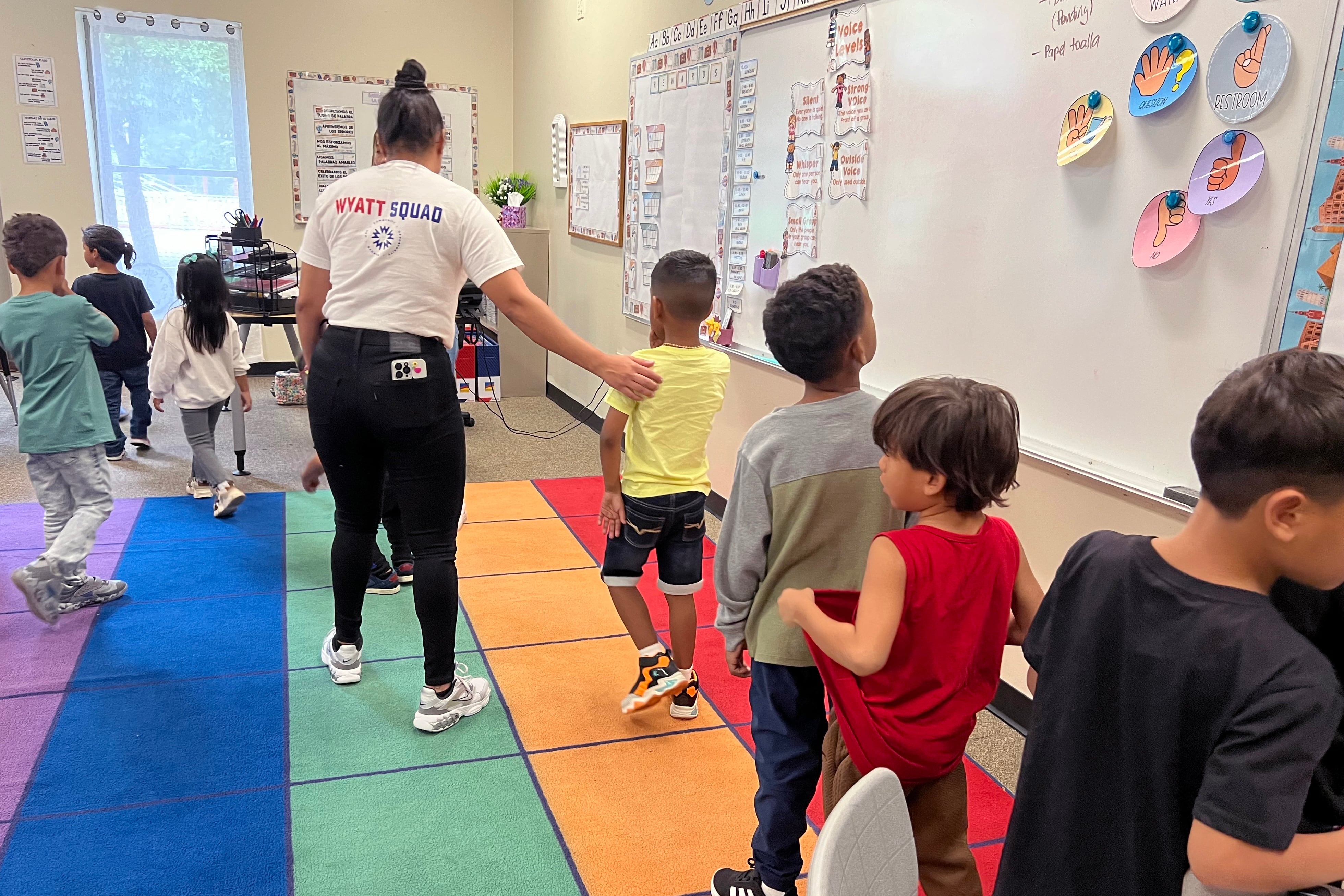 An adult leads a line of young students over a colorful rug in a classroom.
