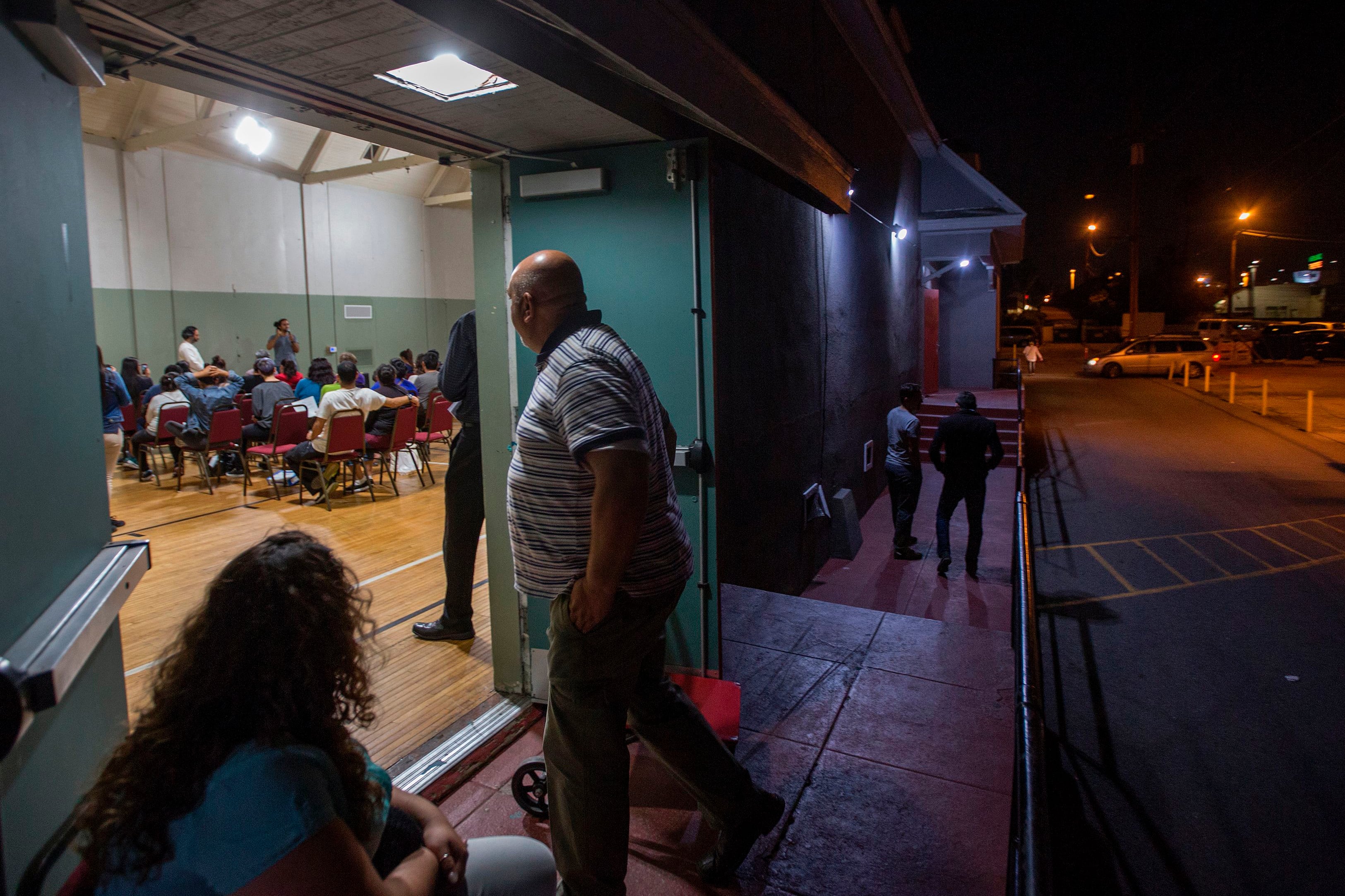 An adult and a child stand in the doorway of a building with people inside and a couple of people standing outside.