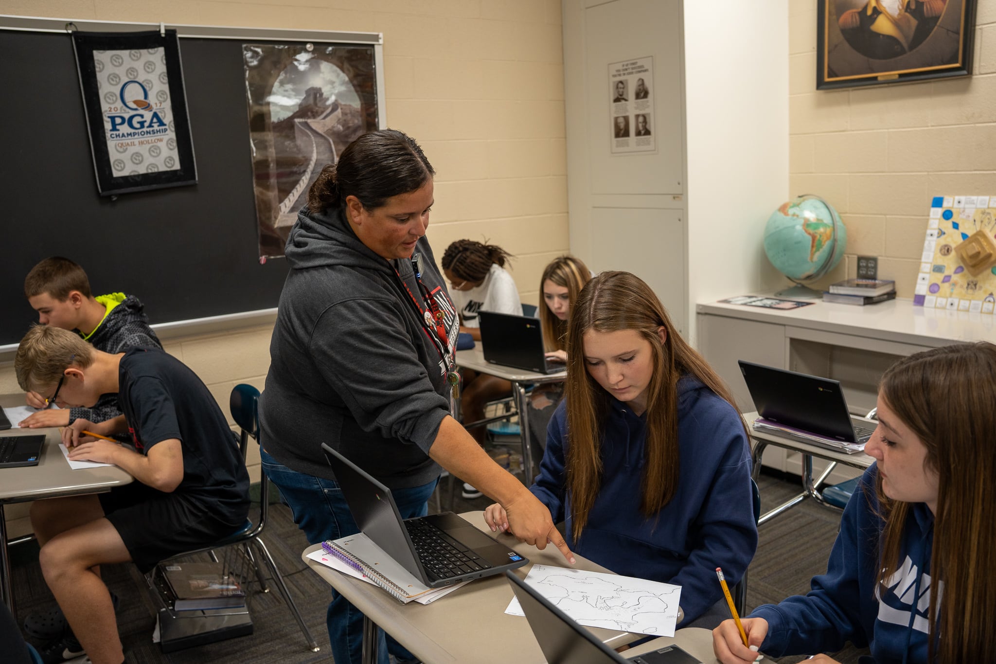 A teacher with brown hair wears a gray sweatshirt. She talks to high school students in her classroom.