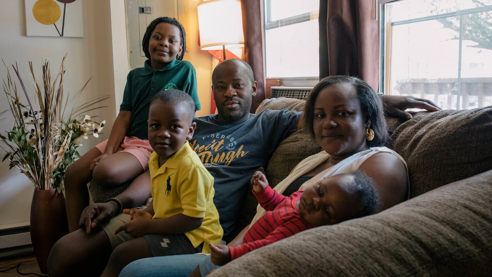 The Dairo family, consisting of parents John and Oyefunmilola and their young children, Abigail, Michael and infant Elijah, sit together on their living room couch next to a window.