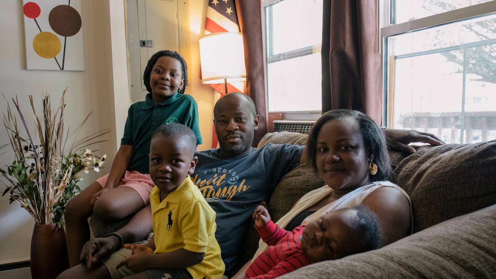 The Dairo family, consisting of parents John and Oyefunmilola and their young children, Abigail, Michael and infant Elijah, sit together on their living room couch next to a window.