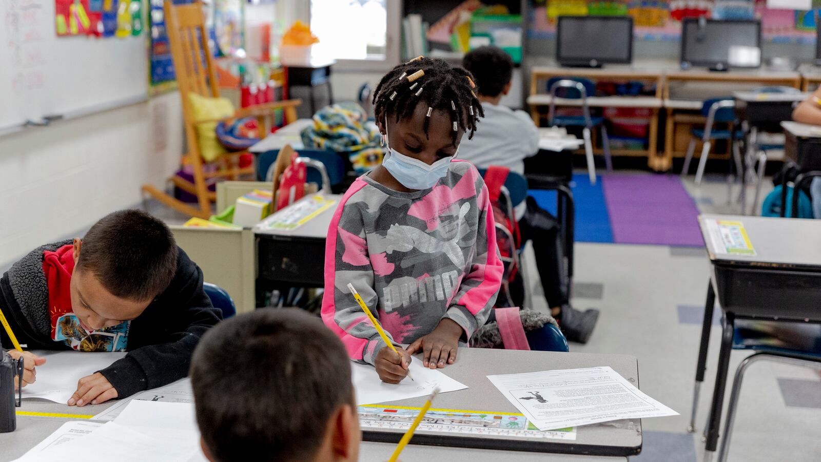 A child with braids and wearing a mask and a pink and gray sweater writes on a piece of paper with a pencil while standing at a desk