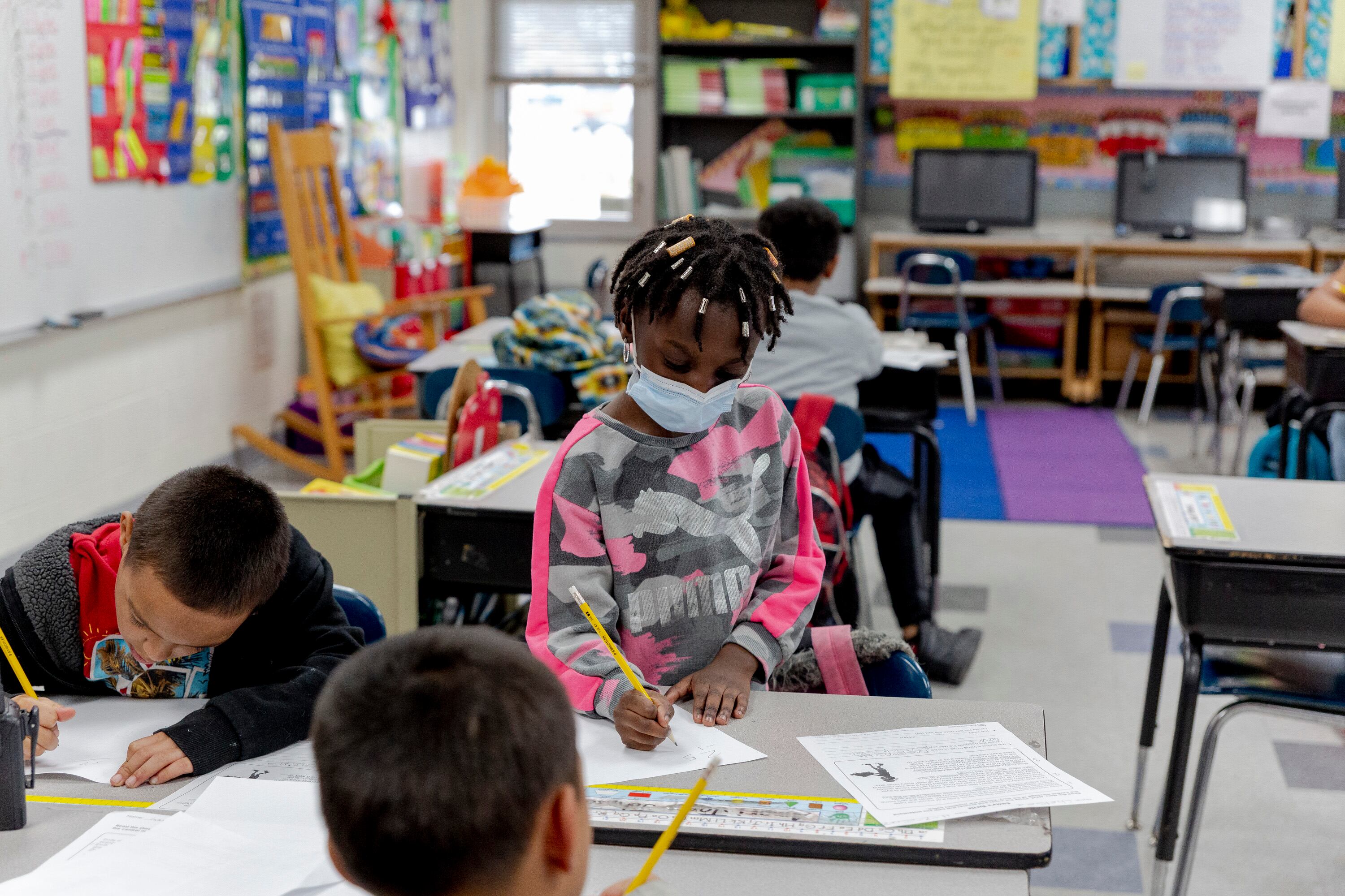 A child with braids and wearing a mask and a pink and gray sweater writes on a piece of paper with a pencil while standing at a desk