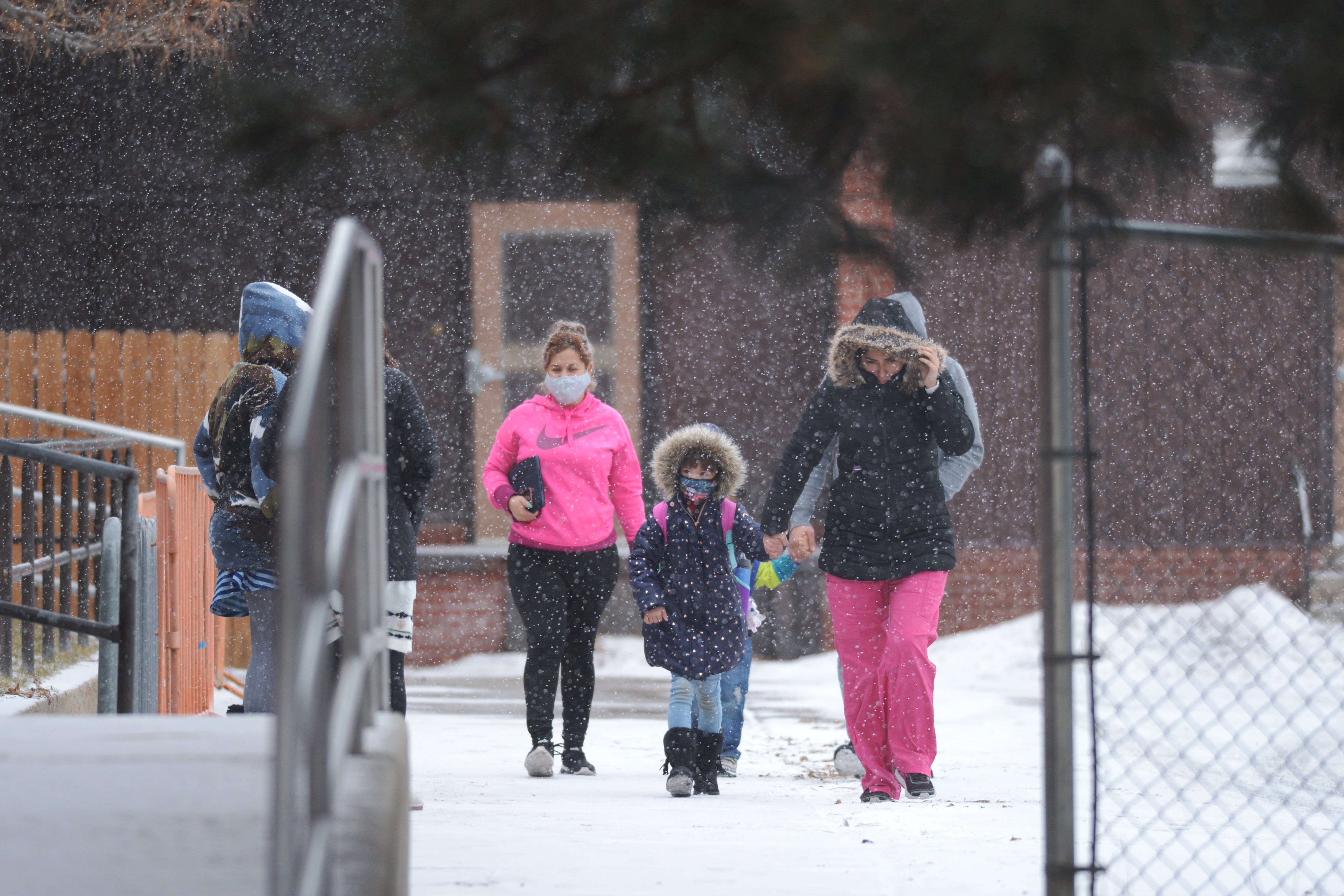 Parents and children wearing winter coats and masks walk to school on a snowy day.