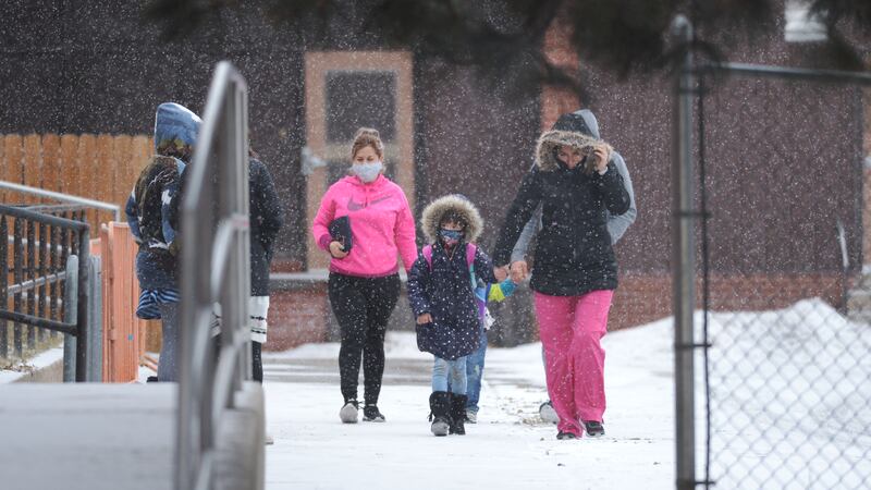Parents and children wearing winter coats and masks walk to school on a snowy day.