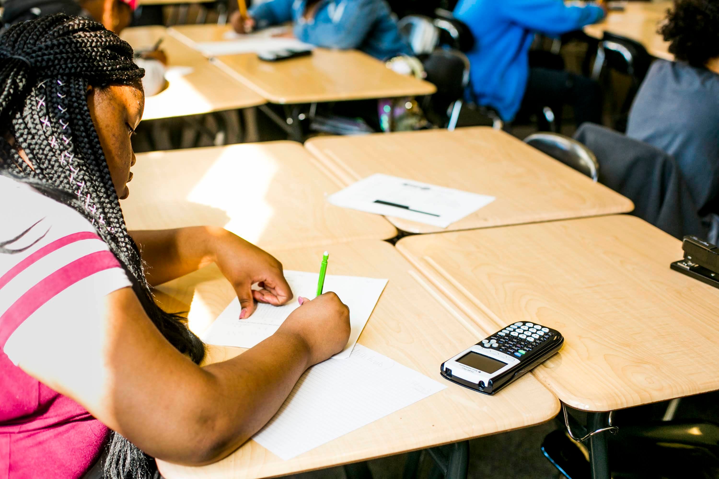 A girl at a student desk writing on a piece of paper. A calculator is also on the desk.