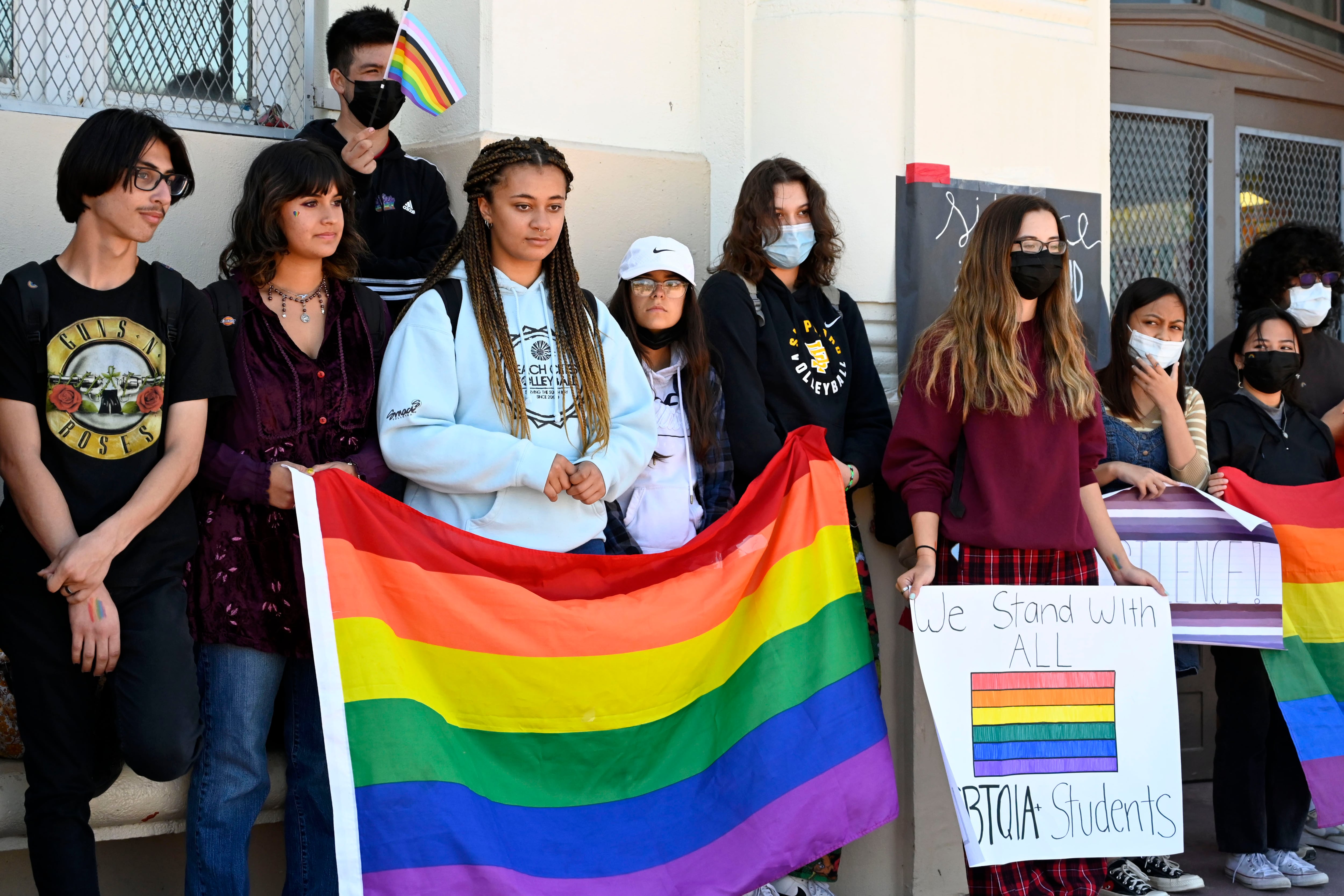 Students protest in support of the LGBTQIA community outside of their school, holding Pride flags and signs.