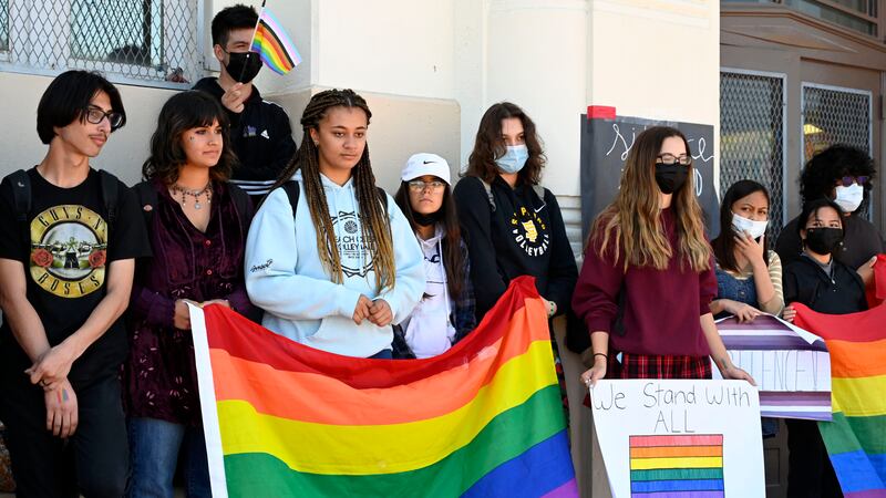 Students protest in support of the LGBTQIA community outside of their school, holding Pride flags and signs.