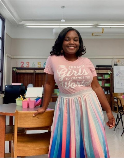 Photo of a young, Black female teacher wearing a pink shirt that says, "Educate girls to change the world" and a multicolored skirt.