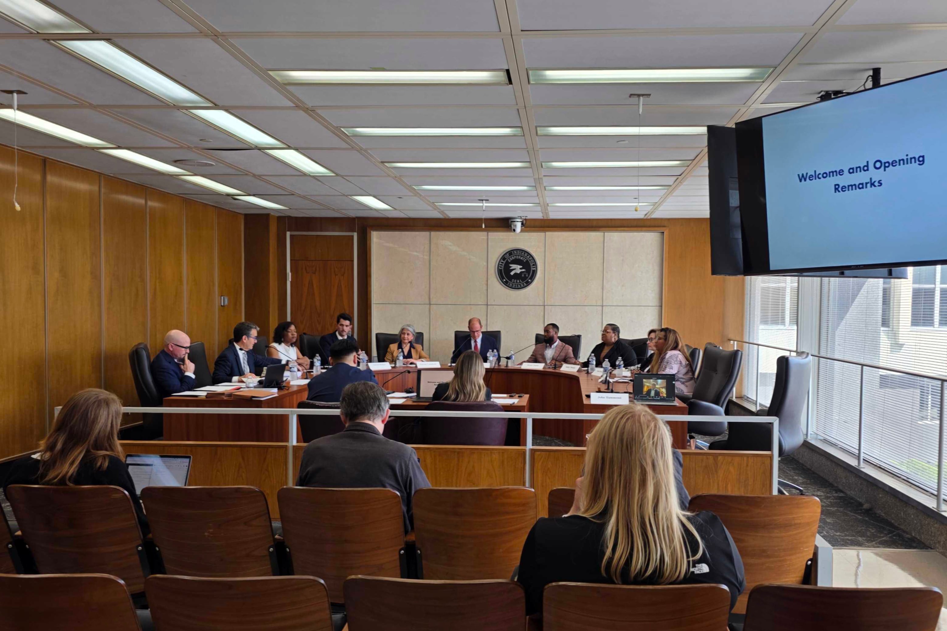 A photograph of a 10 or more adults sitting around wooden tables with people sitting in rows of chairs looking at them and a large TV screen on the right side of the image.