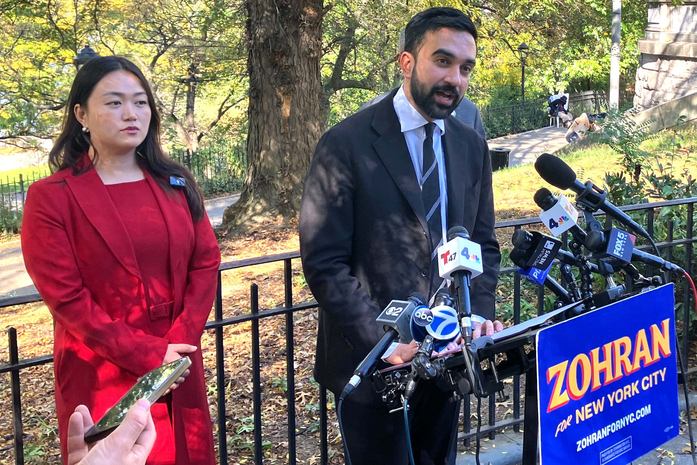 A man in a dark suit speaks behind a bank of microphones.