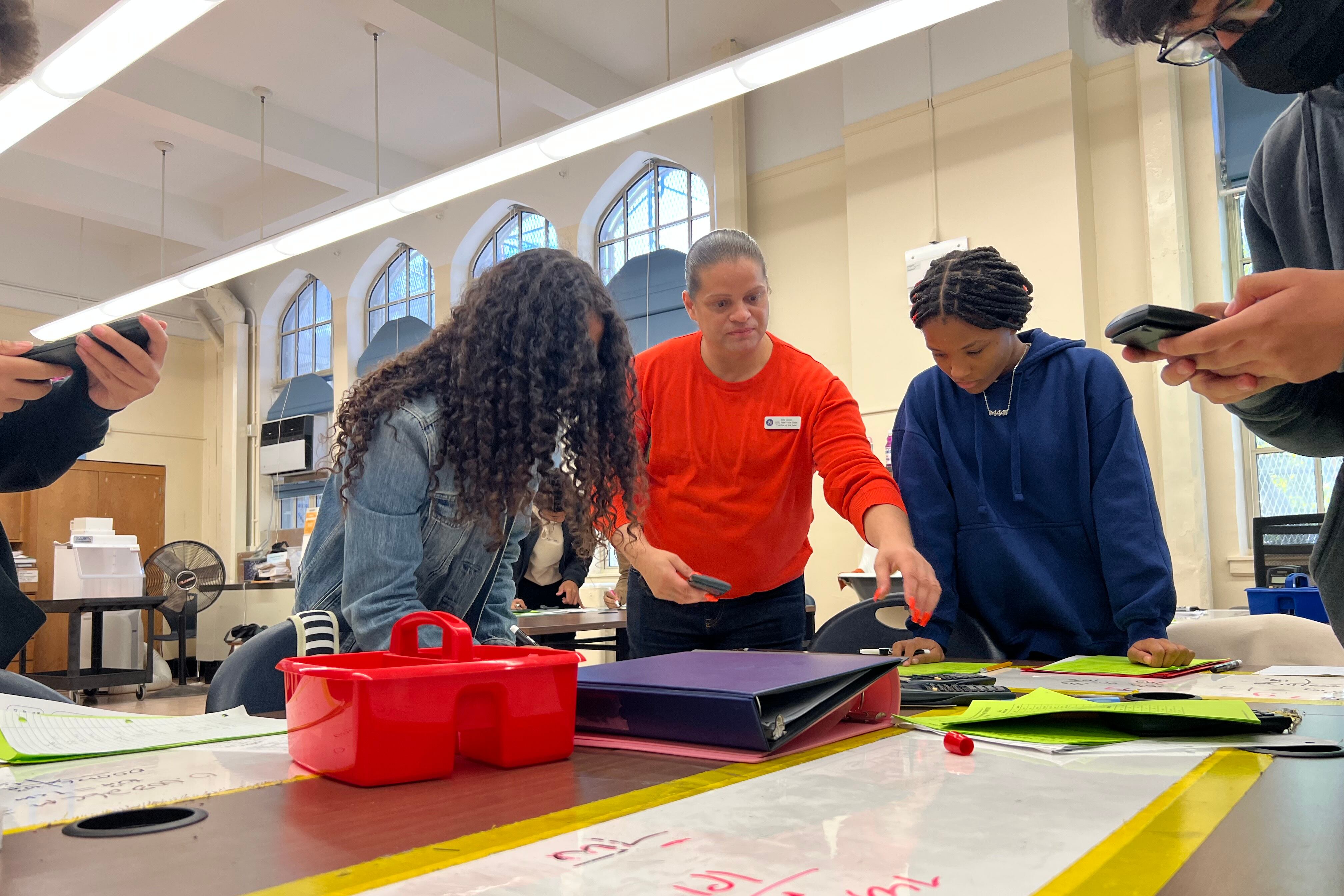 A man in a red shirt gestures toward a table with two students on either side of him.