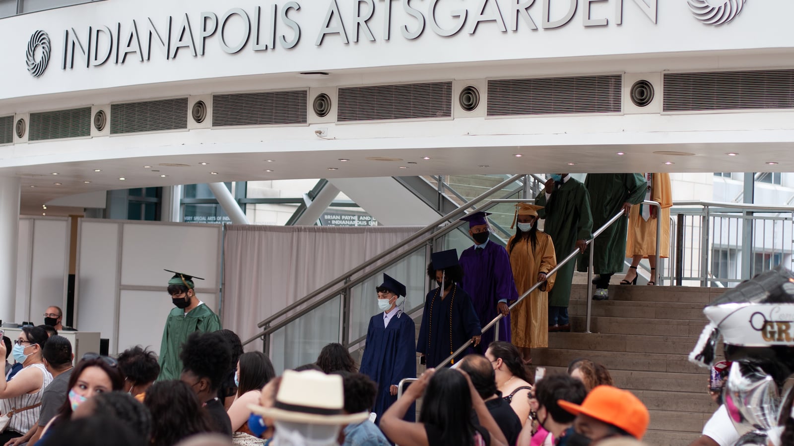 Graduating students, wearing traditional garb, walk down a set of stairs underneath a large structure that reads “Indianapolis Artsgarden” as family look on.