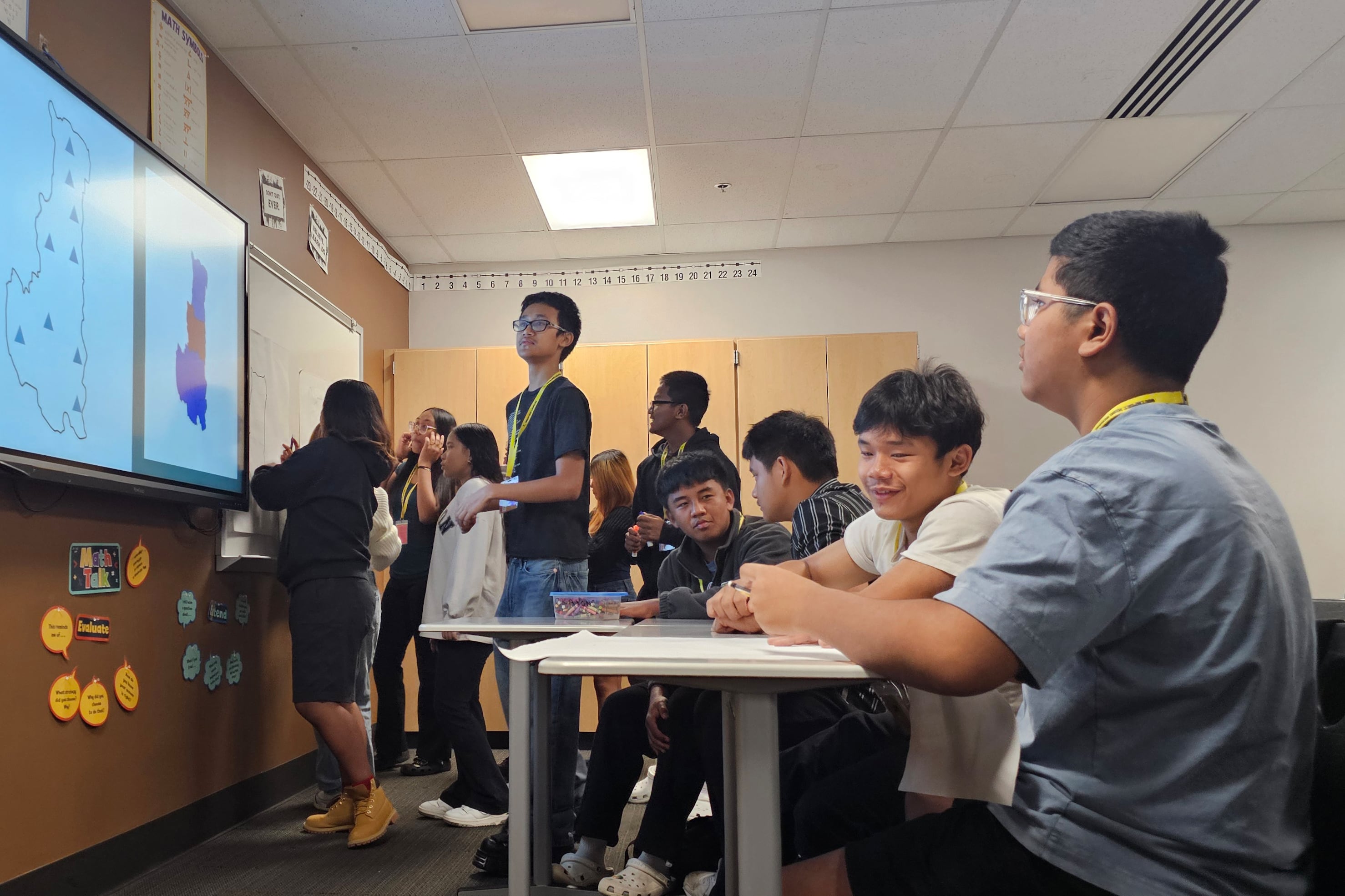 A large group of students in a classroom - some are sitting at tables and others are standing.