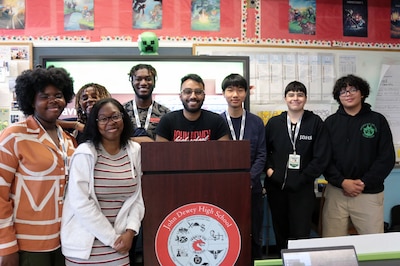 Students and a teacher pose for a photograph in front of a dry erase board and behind a wooden podium in a classroom.