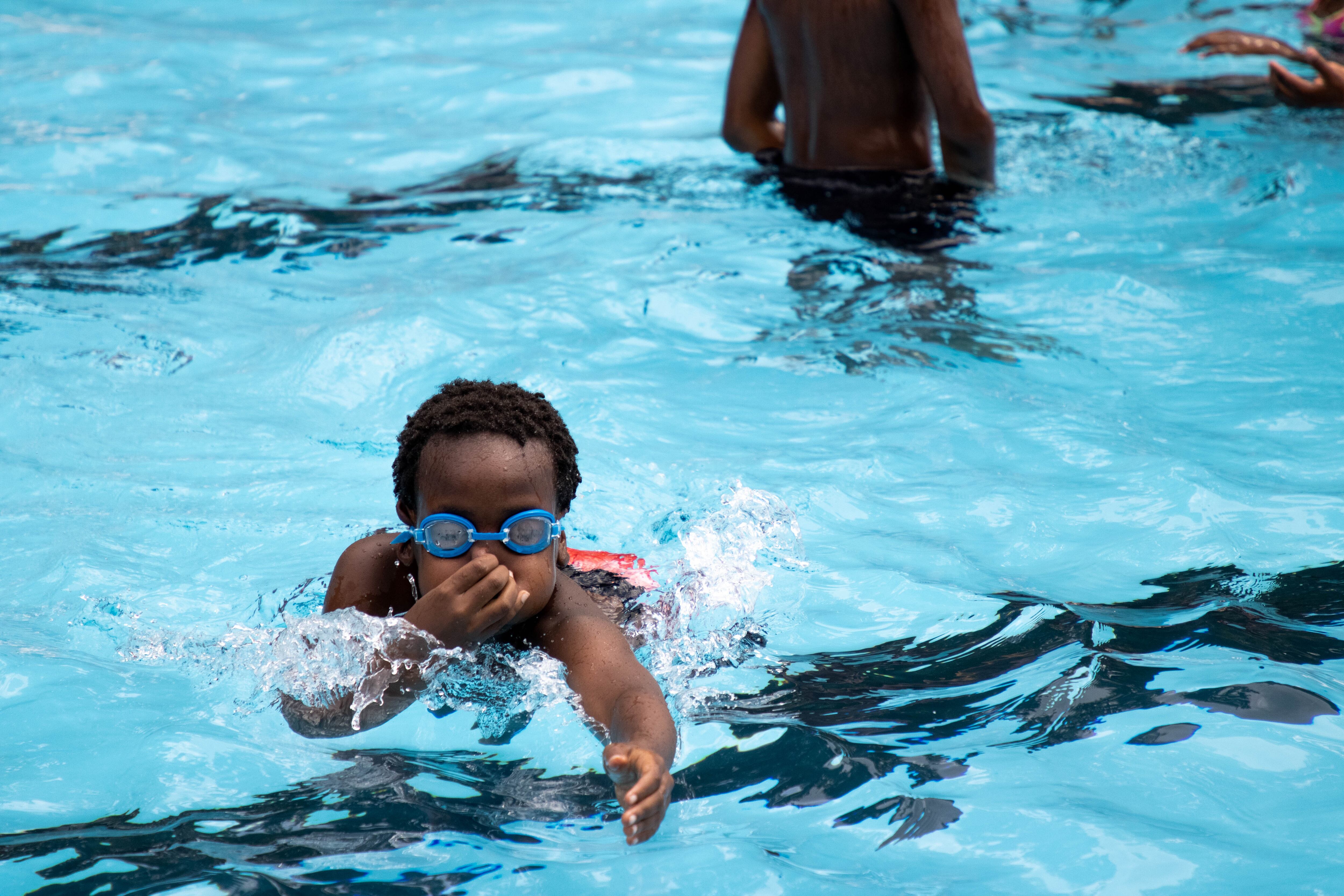 A child with blue goggles holds his nose as he swims in an outdoor pool.