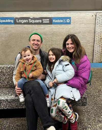Two adults hold two young kids and all smile and look at the camera while sitting on a stone bench at a train station.