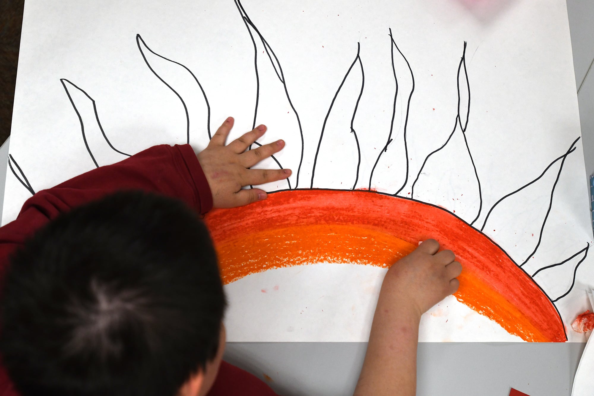 A dark-haired child is seen from above working on an art project. A half circle and rays depict a sun. The child is coloring a long orange arc.
