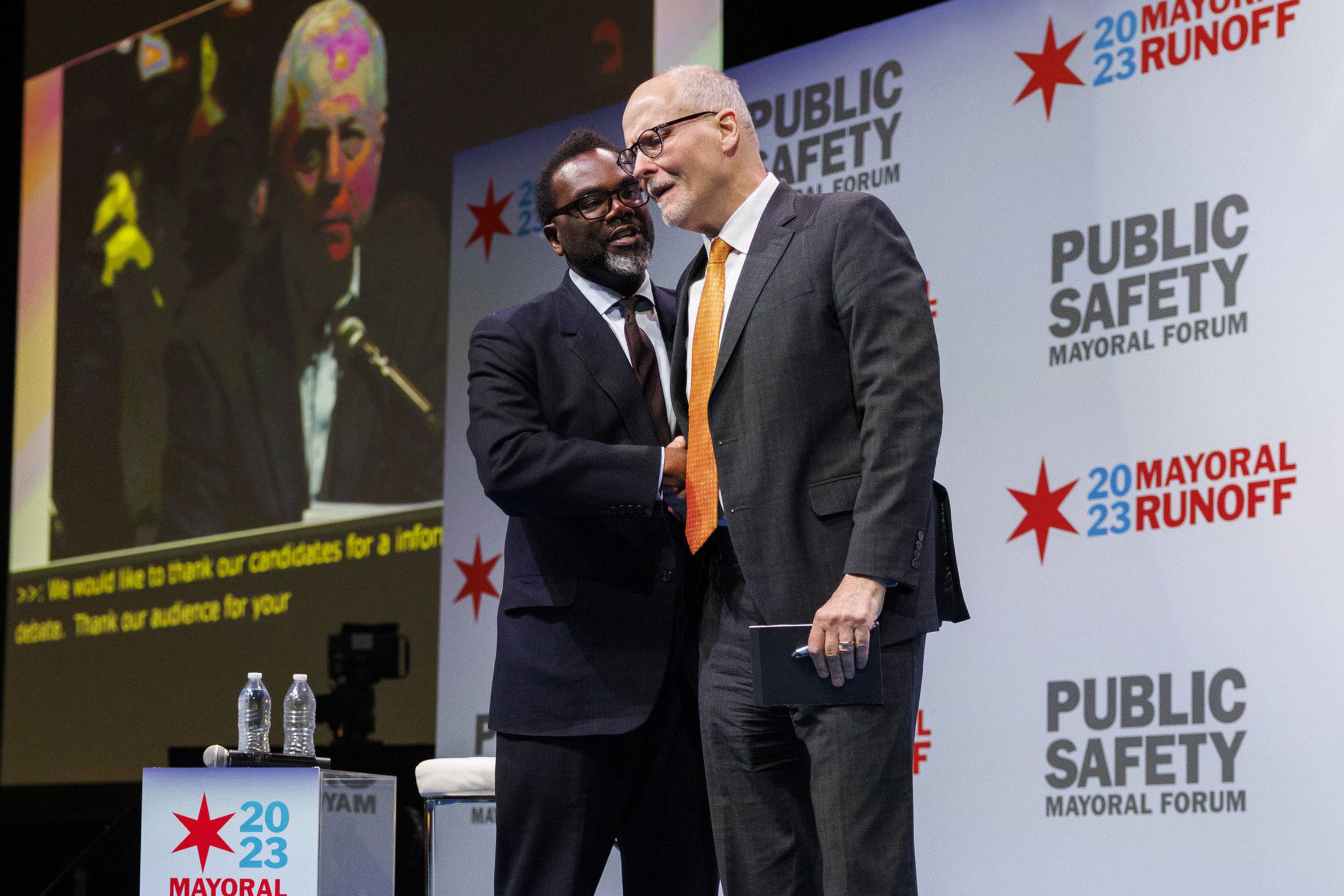 Two men in suits and ties stand close together on a stage, in front of a screen showing a man with a microphone.