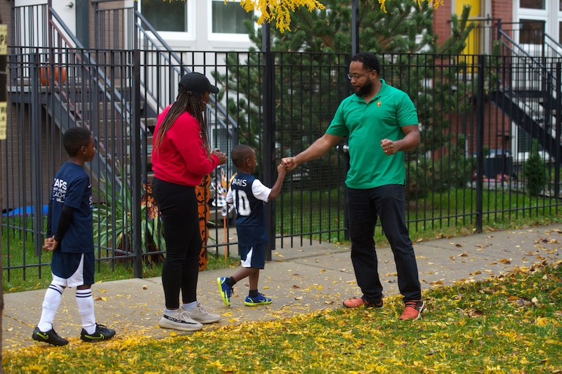 A man wearing a green shirt greets two young students and an adult outside.