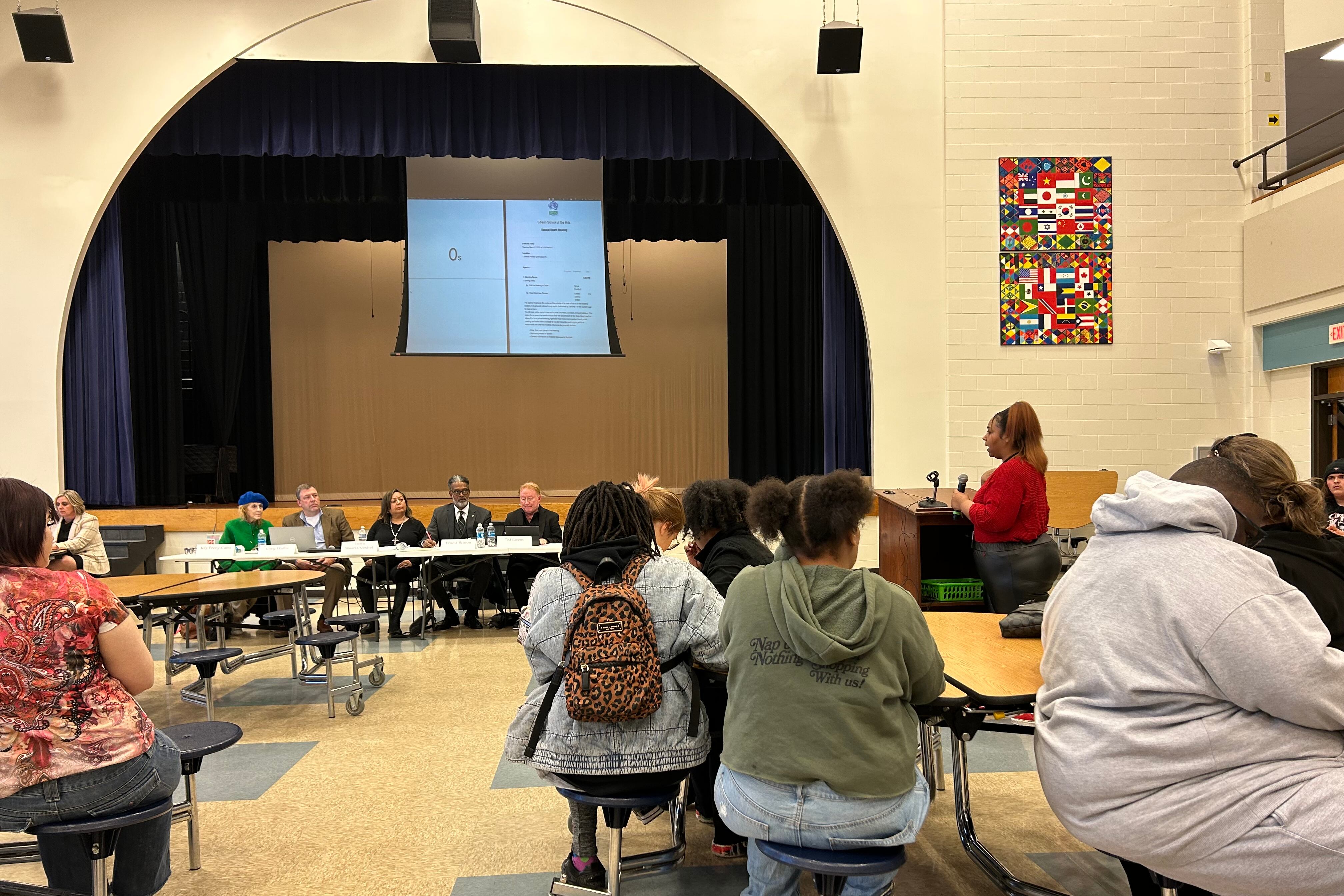 A woman in a red shirt at a podium addresses a row of people sitting behind a table in a school cafeteria. In the foreground are other people sitting and listening to her comments.
