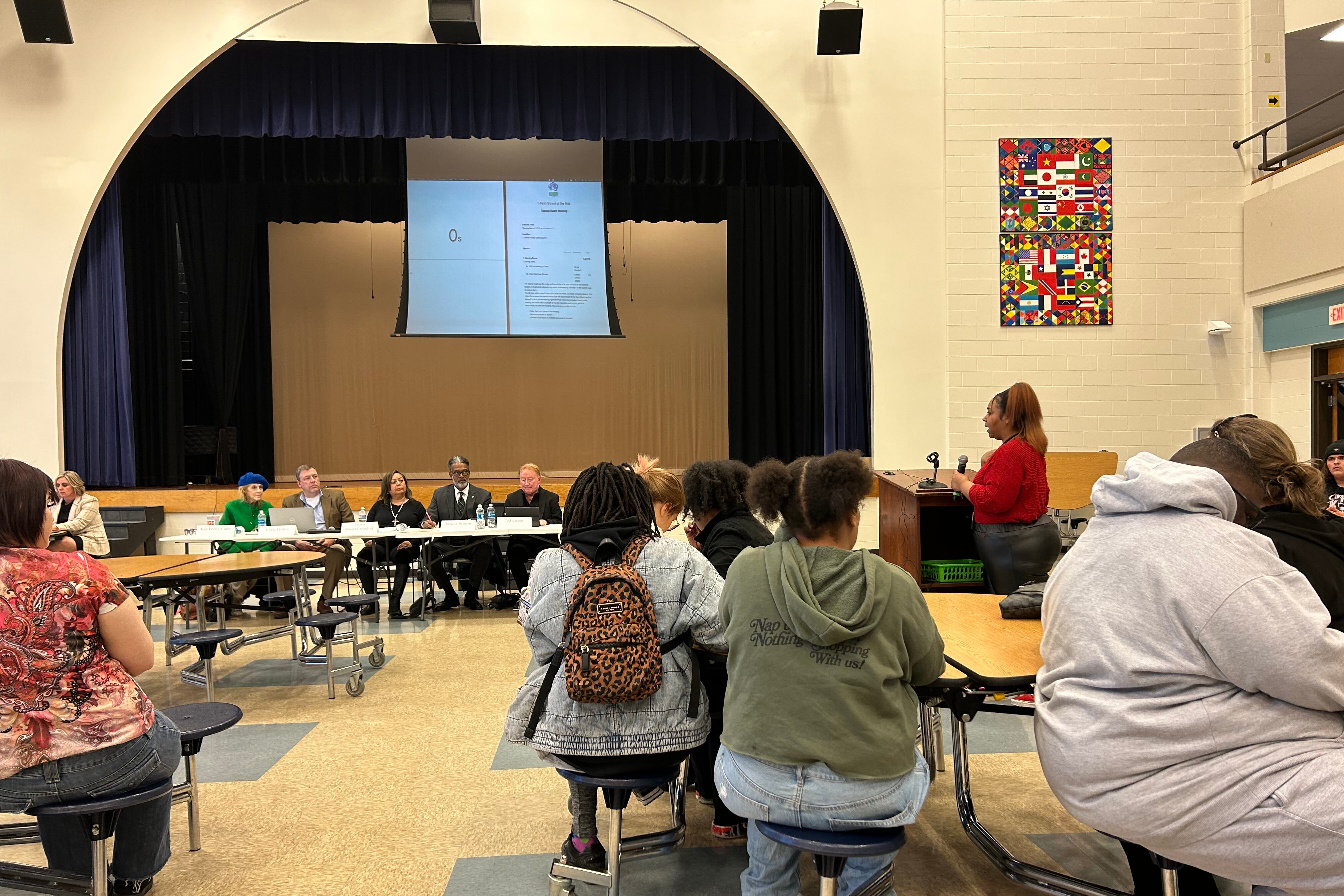 A woman in a red shirt at a podium addresses a row of people sitting behind a table in a school cafeteria. In the foreground are other people sitting and listening to her comments.