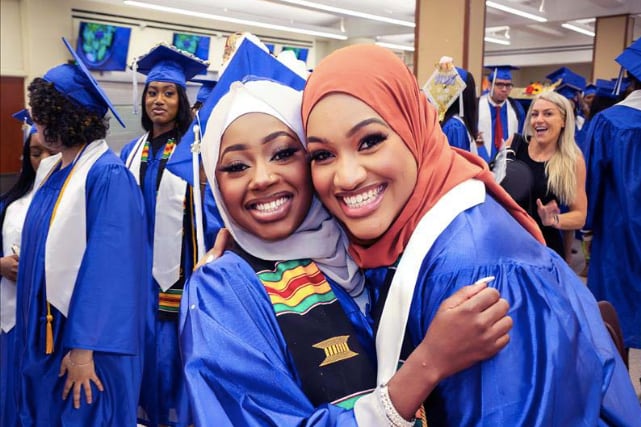 Two high school graduates in hijabs and caps-and-gowns smile at in a crowded room for a graduation ceremony.