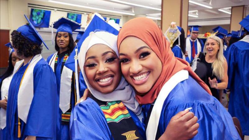 Two high school graduates in hijabs and caps-and-gowns smile at in a crowded room for a graduation ceremony.