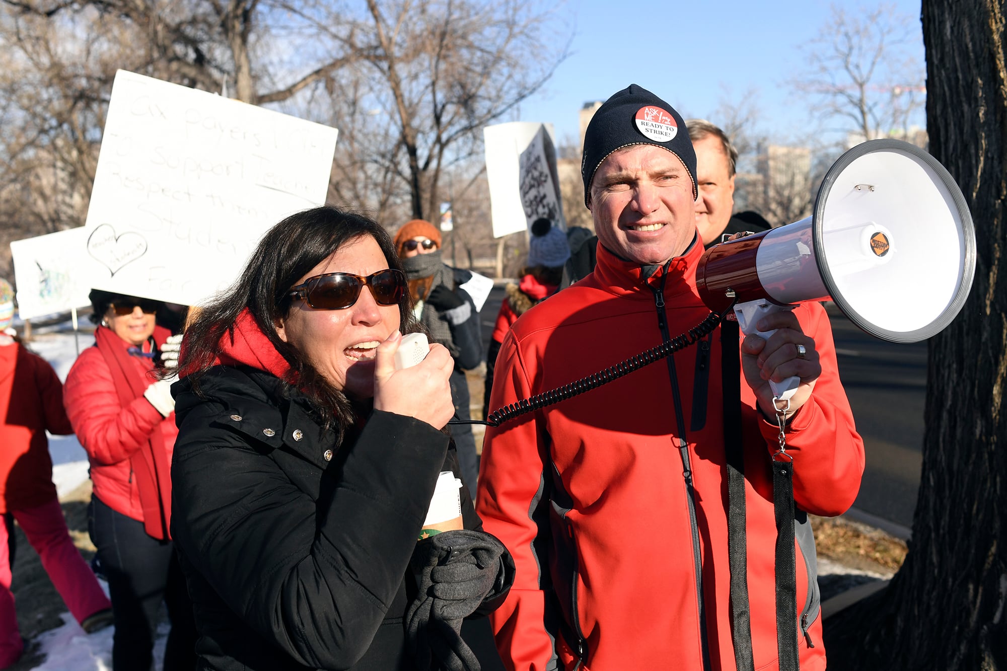 Two people in coats hold up a microphone outside next to a group of protestors.