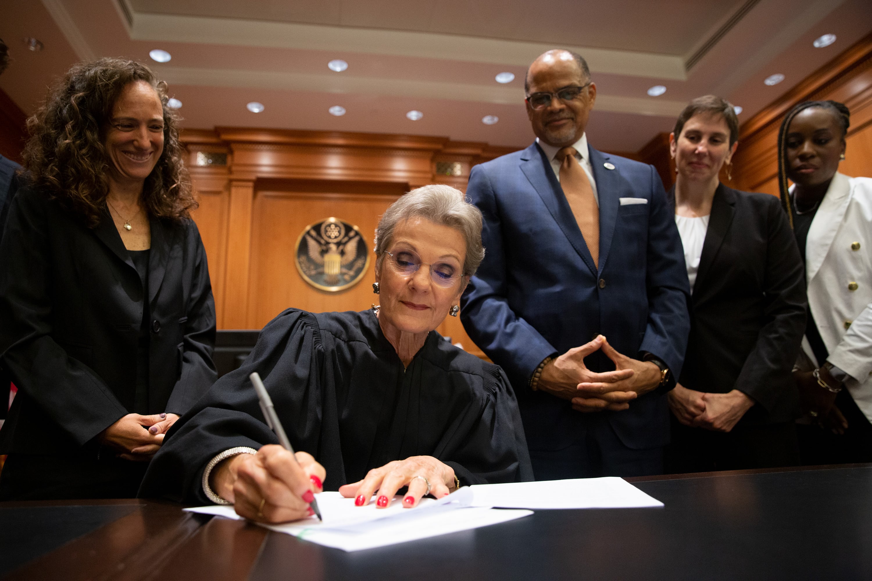 A female judge in robes sits at a table and signs a court order flanked by other officials.