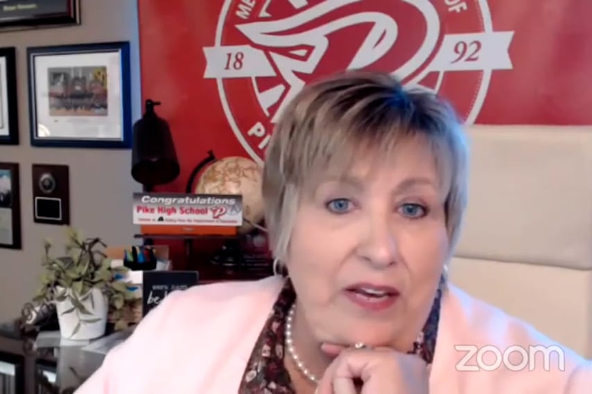 In a screenshot of a video, a woman is speaking to the camera with her chin resting in her hand. The office wall behind her is decorated with a red school logo, picture frames, and plaques.