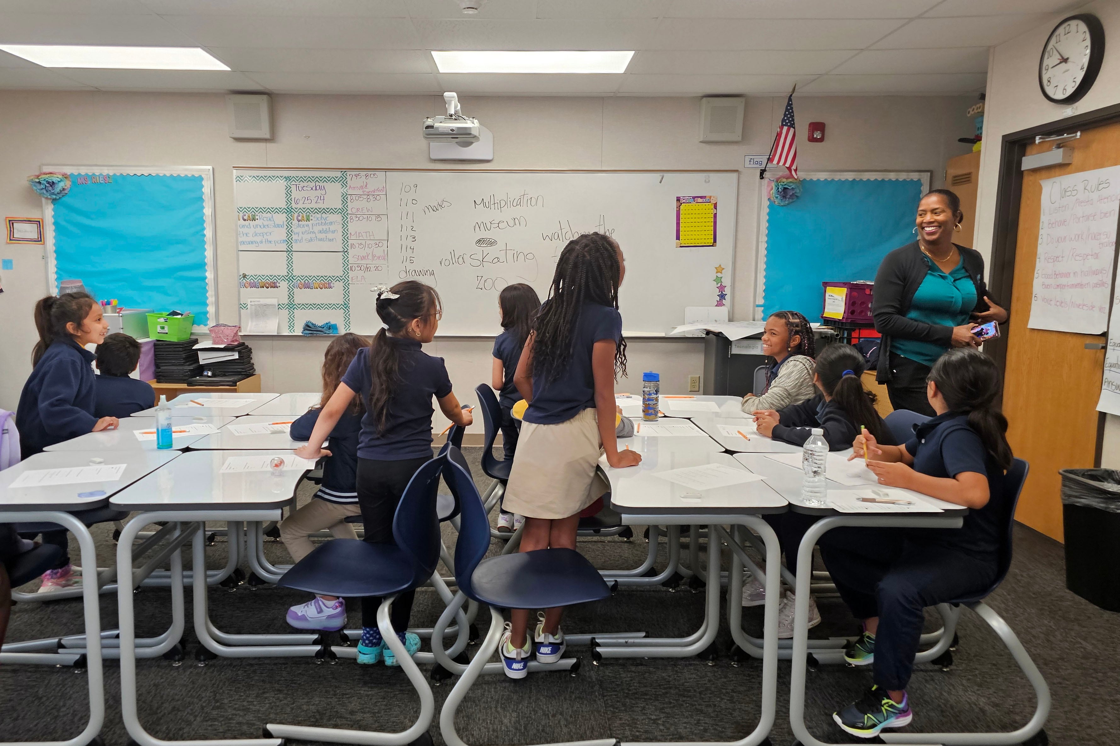 An adult stands toward the back of a classroom with students sitting at two tables.