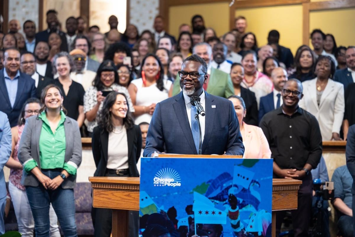 A man in a suit and tie smiles at a lectern while other people stand behind him.