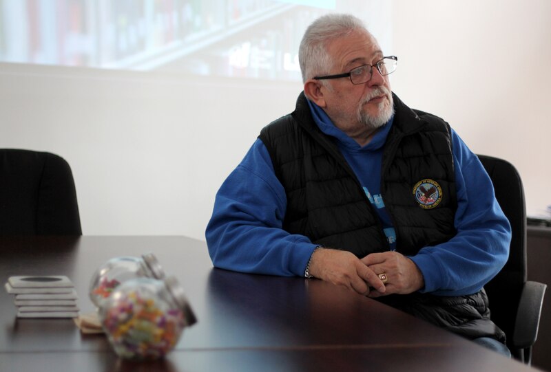 A man wearing glasses and a black vest sits at a table with a projected image in the background.