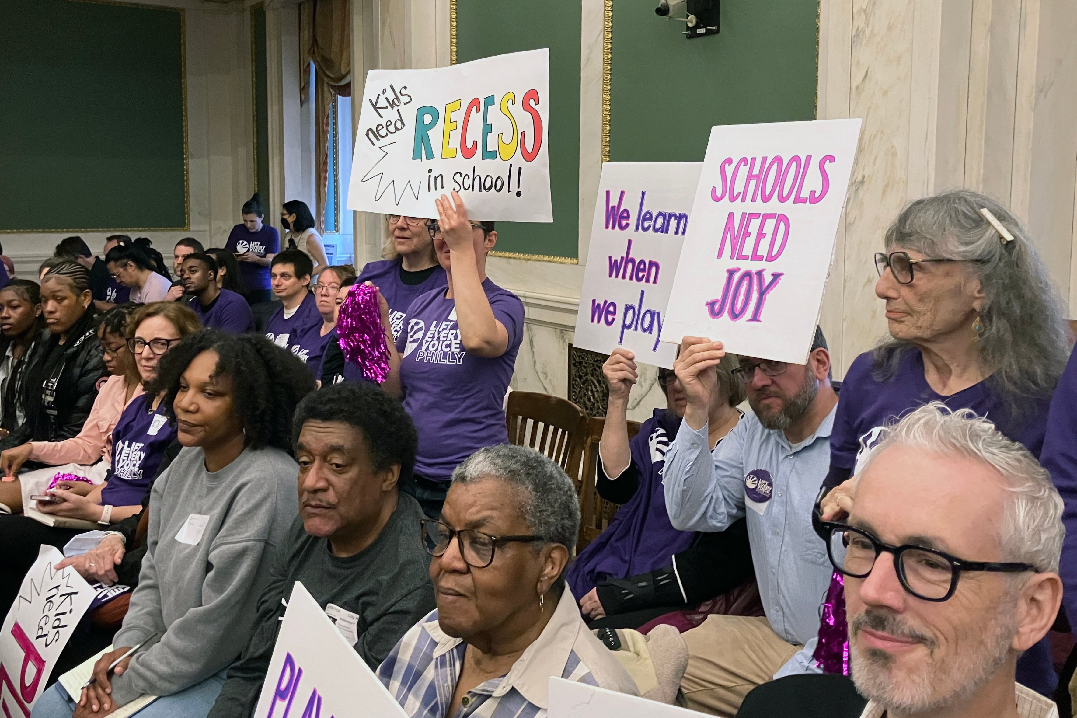 Adults and students gather together in a corner of a large meeting room, some are holding signs.