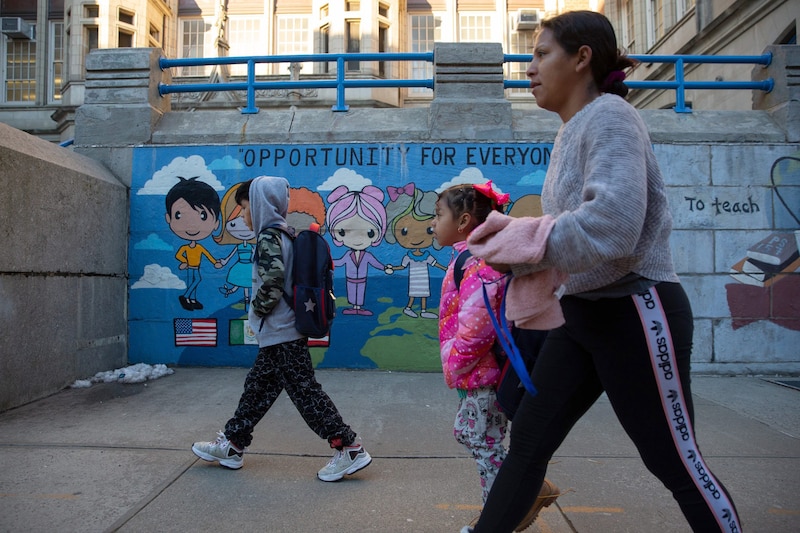 Two young students and an adult walk outside with a mural on a wall in the background.