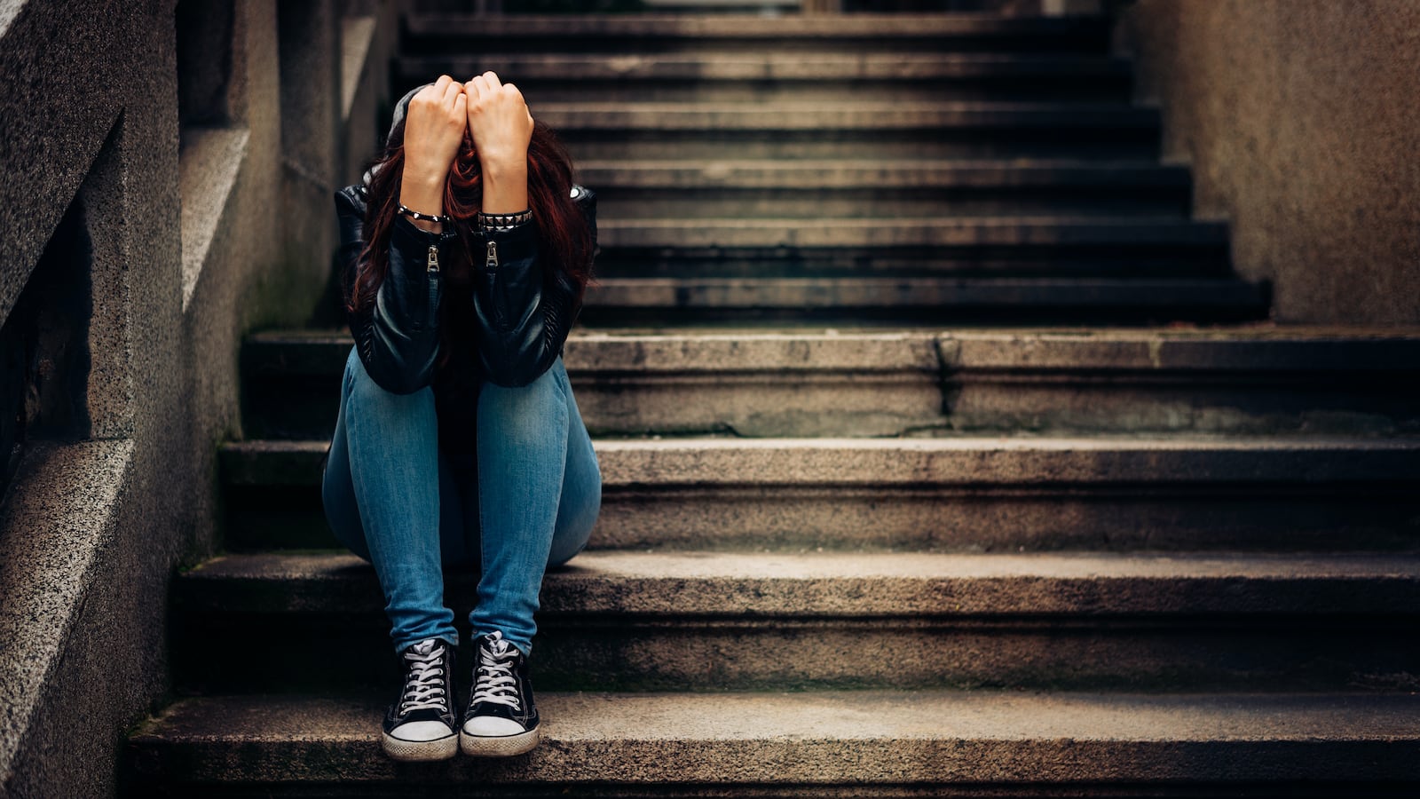 A young person wearing jeans and Converse-style shoes sits on concrete steps holding their head in their hands