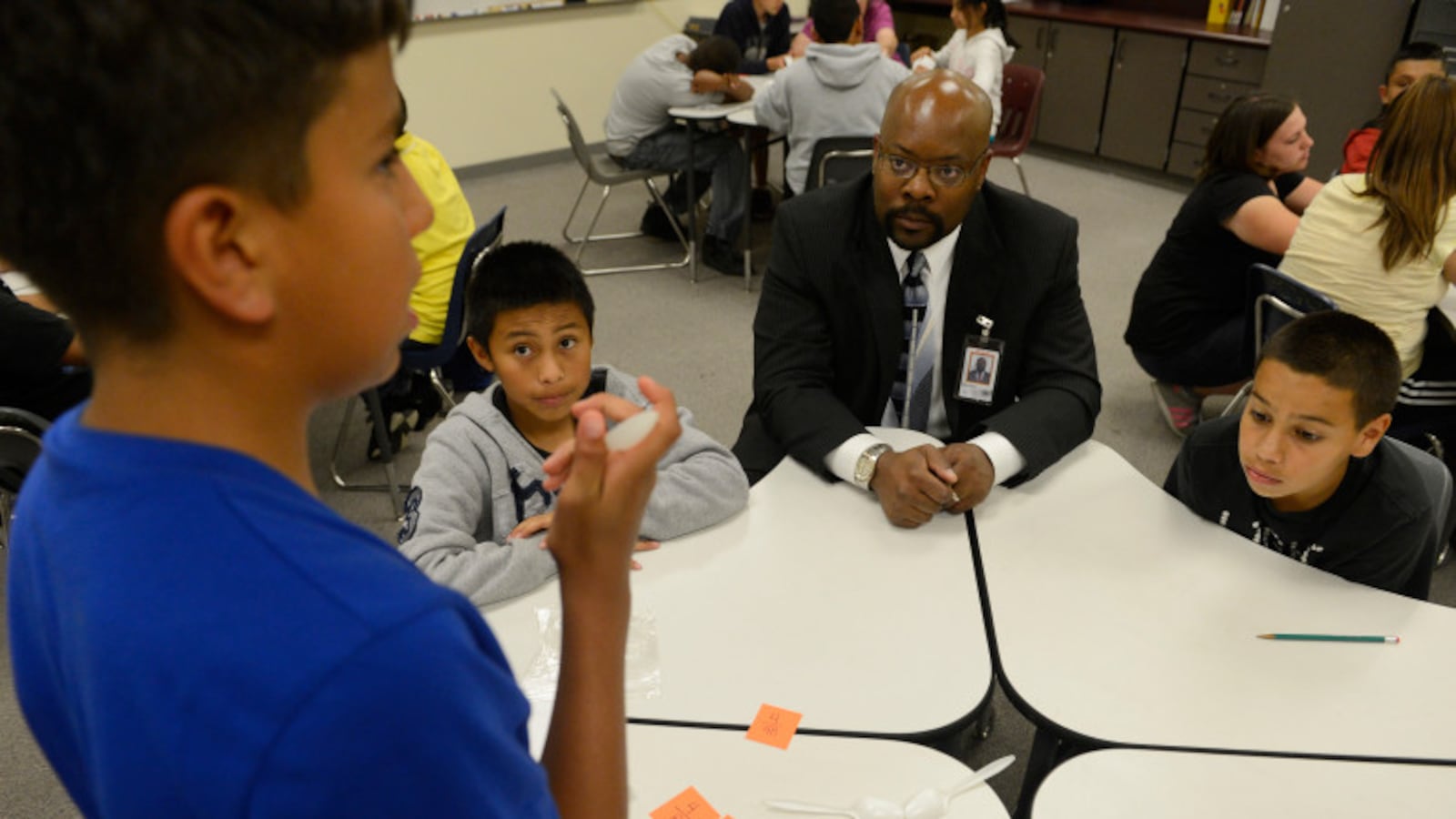 A boy in a blue shirt works on a math card game problem with two other students seen at the table while Superintendent Rico Munn sits on the corner of the table, wearing a black suit and tie.