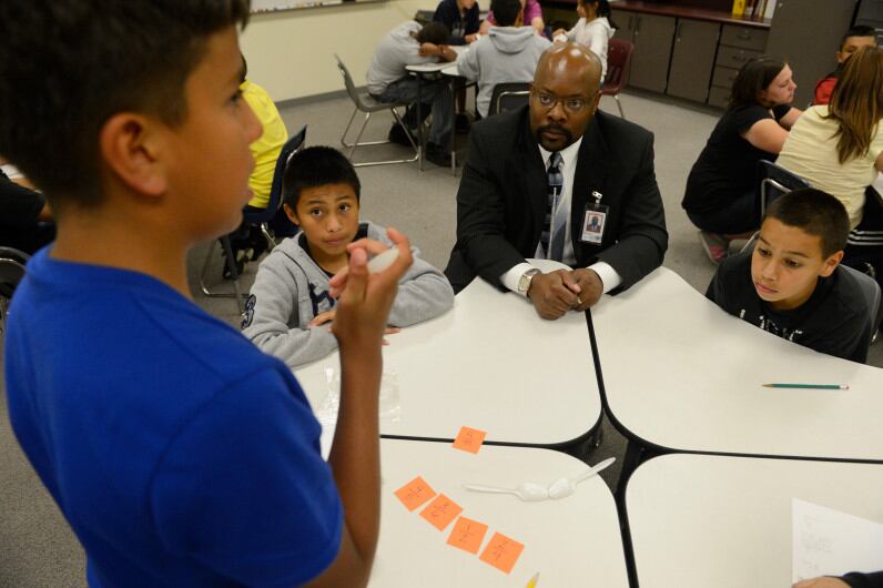 A boy in a blue shirt works on a math card game problem with two other students seen at the table while Superintendent Rico Munn sits on the corner of the table, wearing a black suit and tie.