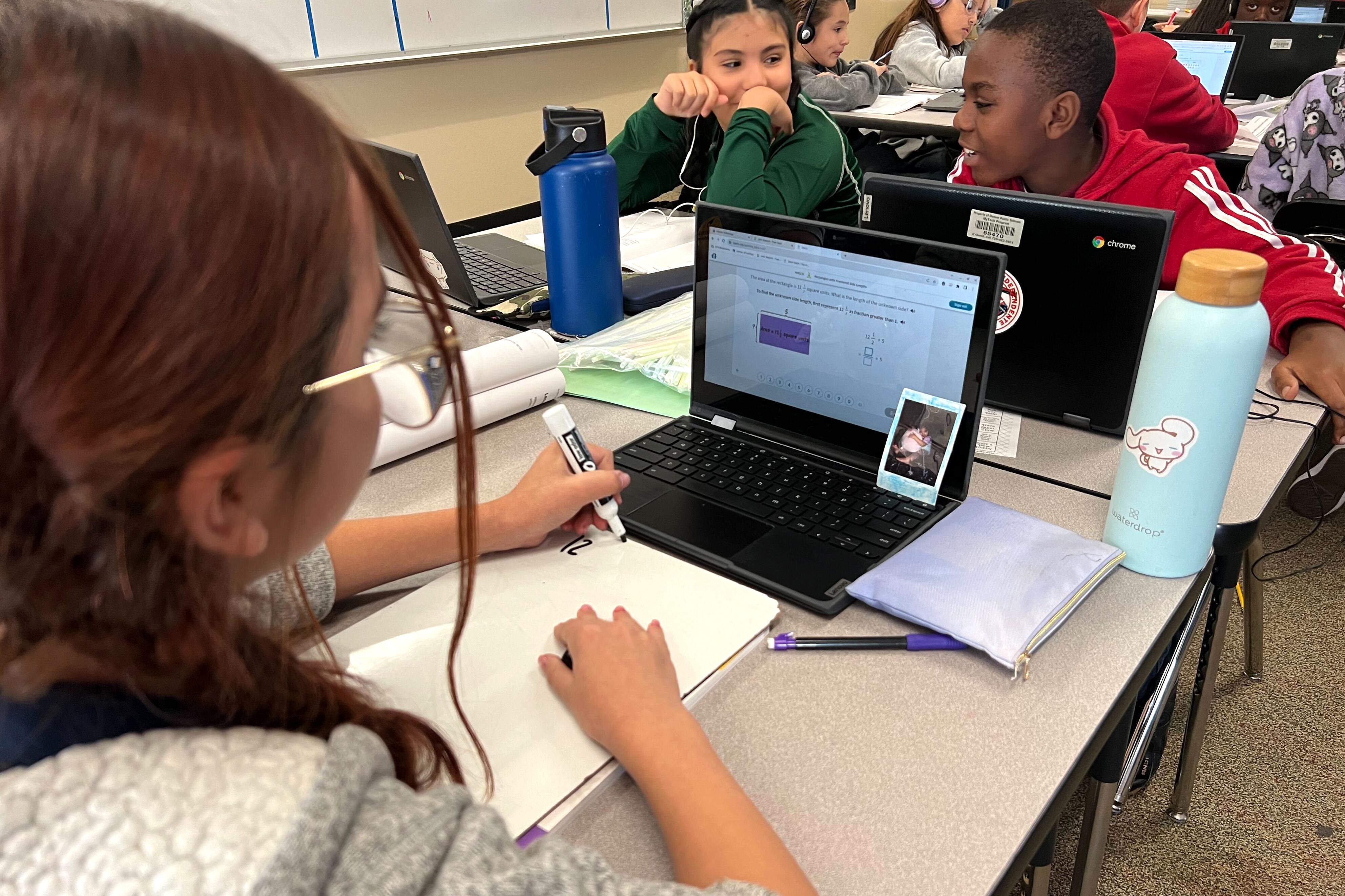 Three students work at their desks while other students work in the background.